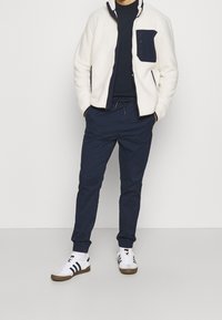 Fleecejacka i vitt med en mörkblå dragkedjeficka, matchad med marinblå byxor och vita sneakers med marinblå och bruna detaljer.