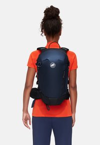 Zaino blu navy con una texture liscia e lucida, dotato di spallacci regolabili, una tasca frontale e un logo sul pannello posteriore.