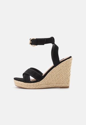 Sorte kilehæle sandaler med en vævet jute platform, justerbar ankelstrop og krydsdesign på overdelen med kontrasterende stikninger.