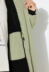 Olijfgroene jas met zwarte fleecepanelen; heeft een ritszak met een neongele ritsgaatje; gestructureerde stof en een gladde voering.