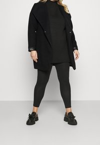 Mustat takki leveällä kauluksella, tumman yläosan päällä, yhdistetty mustiin legginsseihin ja paksupohjaisiin kenkiin, joissa on nauha. Pintakankaat ovat sileitä.