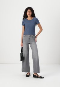 Blå ribbad t-shirt med kort ärm, grå vida jeans, svarta platta skor med ögla-detaljer och en liten svart handväska. Enformig vit bakgrund.