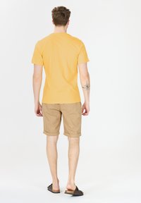 Gul bomullströja, tan shorts med uppvikta fållar och bruna slip-on sandaler. Enkelt mönster med en avslappnad passform, sedd från baksidan.