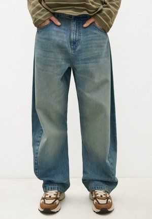 Personne portant un jean bleu deux tons ample avec les mains dans les poches, associé à des baskets marron et blanches et un t-shirt vert à rayures à manches longues.