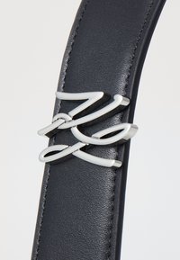 Svart lærbelte med en glatt tekstur, med et sølvmetalllogo i midten. Enkelt, elegant design.