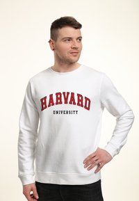 Bílá mikina s nápisem "HARVARD" v červené barvě a "UNIVERSITY" v černé barvě, s vroubkovanými manžetami a lemem, hladkou texturou a designem s kulatým výstřihem.
