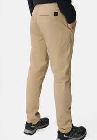 Beige Hosen haben eine schlanke Passform, eine Gesäßtasche und dezente Nähte. Kombiniert mit schwarzen Turnschuhen, die graue und gelbe Akzente haben.