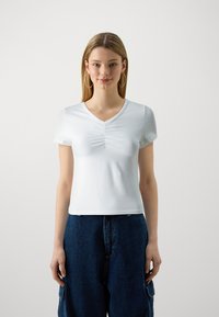 Hvit tettsittende T-skjorte med korte ermer og V-hals, med rynkedetaljer i midten. Båret sammen med mørkeblå baggy denimbukser.