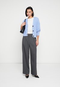 Ανοιχτό γαλάζιο πλεκτό ζακέτα με μεγάλα κουμπιά, λευκό εφαρμοστό τοπ, γκρι παντελόνι με φαρδιά πόδια και μαύρα παπούτσια με μυτερή μύτη, κρατώντας μια μαύρη τσάντα.