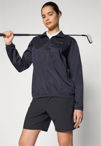Marineblauwe quarter-zip windbreaker met elastische zoom, gecombineerd met zwarte shorts. Heeft een kraag en een logo op de borst. Gladde textuur.