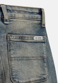 Jeans in denim di colore azzurro chiaro con un lavaggio sbiadito. Presentano una tasca posteriore con etichetta bianca "GUESS" e cuciture a contrasto. Vestibilità standard.