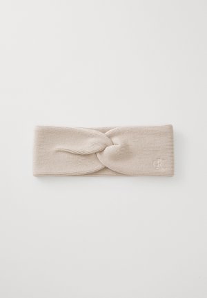 Knit headband in light beige, featuring a twisted design at the front and embroidered logo on the right side. Soft, textured fabric.