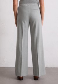 Pantaloni grigi chiari a gamba larga realizzati in un tessuto liscio e strutturato, dotati di una tasca posteriore e di una comoda vita elastica. Indossa scarpe marroni.