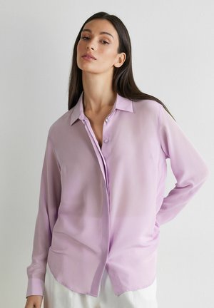 Kvinde med langt mørkt hår iført en lys lilla skjorte med knapper og hvide bukser, stående foran en ensfarvet hvid baggrund.