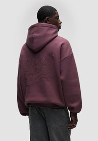 Felpa viola con cappuccio dal taglio rilassato, dotata di testo ricamato tono su tono sul retro e polsini a coste. Abbinata a jeans grigi scuri.