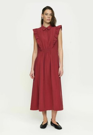 Mladá žena stojaca, oblečená v tmavočervenom midi šatách bez rukávov s volánikovým lemom a čiernymi plochými topánkami, neutrálne svetlé pozadie.