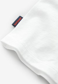 Baltas kokvilnas t-krekls stūrī, kurā redzams tumši zils auduma birkas nozīmītē "SUPERDRY" oranžā krāsā. Redzama maiga tekstūra un noapaļots apakšmala dizains.