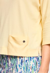 Light yellow top with a pocket and decorative hardware. Features short sleeves and a relaxed fit. Paired with patterned green and blue bottoms.