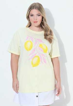 Kobieta ubrana w jasnżółtą koszulkę z różowymi liśćmi i wzorem trzech żółtych cytryn, w połączeniu z białą spódnicą zapinaną na guziki.