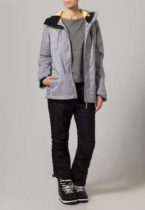 Femme portant une veste gris clair, un pull gris, un pantalon noir et des baskets noires à lacets blancs, debout devant un fond uni.