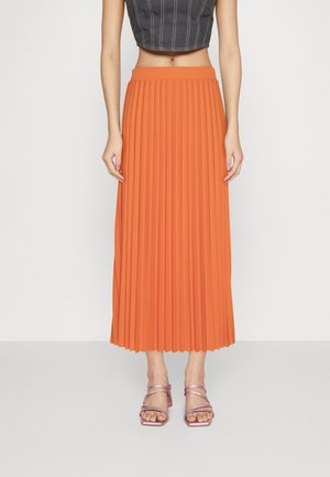 Femme portant une jupe midi plissée orange, un top court noir avec des coutures blanches et des sandales à talons métalliques à brides.