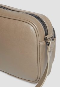 Beige Ledertasche mit glatter Textur, runder Form, silberner Hardware und einem kontrastierenden schwarzen Reißverschluss entlang der oberen Kante.