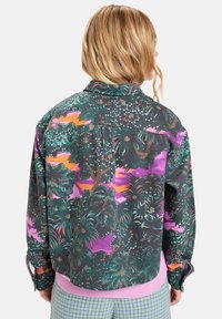 Kragengenielte Jacke aus dunklem Stoff mit einem bunten tropischen Muster von Pflanzen und Bäumen, einem pinken Bund und Knopfmanschetten.