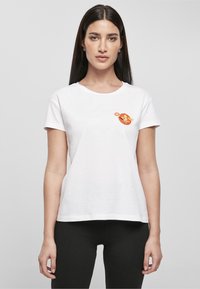 Femme aux longs cheveux foncés porte un t-shirt blanc avec un petit chien de dessin animé dans un anneau orange tenant une balle sur la poitrine en haut à gauche.