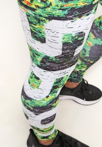 Barevné legíny se zeleným a černým vzorem ve stylu flitrů s bílými akcenty. Tkanina vypadá elasticky; přiléhavý střih. Viditelné černé tenisky.