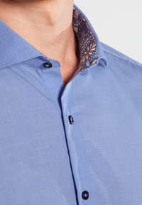 Blue checkered dress shirt with dark buttons and floral patterned inner collar worn by a person, neck visible, on a white background.