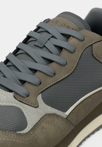 Gray and olive green sneaker featuring suede and mesh materials, textured accents, and flat gray laces. Subtle logo on the tongue.