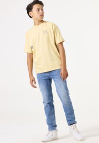 Camiseta de algodón de color amarillo claro con impresiones de texto, combinada con jeans de mezclilla azul claro y zapatillas blancas. El modelo se encuentra en un fondo neutral.