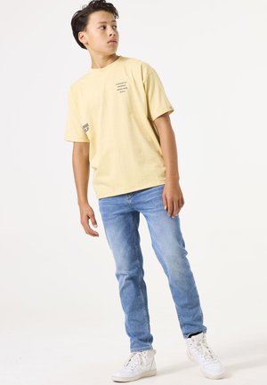 Lichtgele katoenen t-shirt met tekstprints, gecombineerd met lichtblauwe denim jeans en witte sneakers. Het model staat voor een neutrale achtergrond.