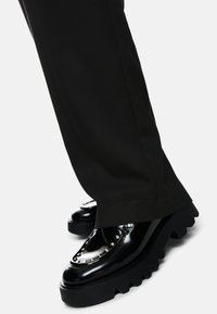 Black chunky platform shoes with silver studs worn with black wide-leg pants on light background.