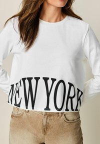 Frau trägt ein weißes Langarm-Crop-Top mit großen schwarzen "NEW YORK"-Buchstaben, kombiniert mit beigen, hoch taillierten Jeans und Ohrgehängern.