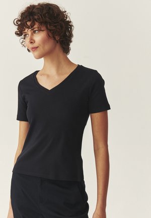 Femeie cu păr creț și scurt, purtând un tricou negru mulat, cu decolteu în V și mâneci scurte, și pantaloni negri, privind spre stânga, pe un fundal simplu.