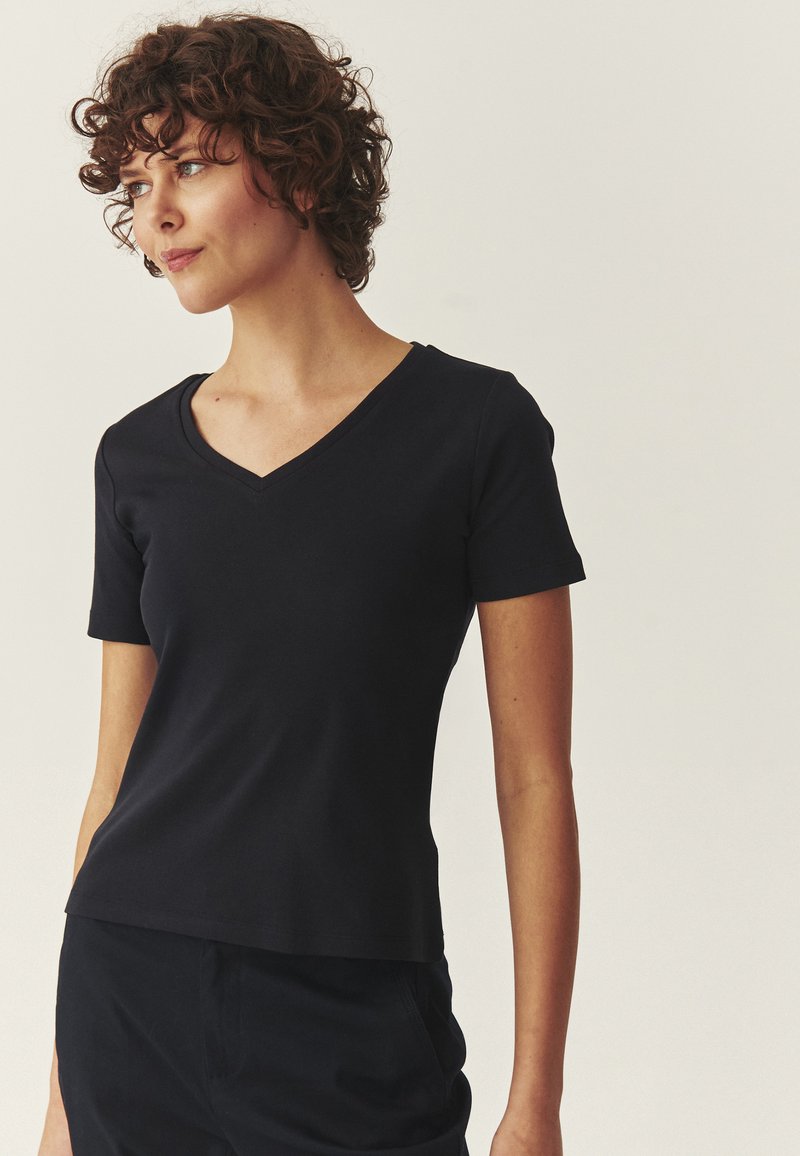 Femme aux cheveux courts et bouclés portant un t-shirt noir ajusté à manches courtes en V et un pantalon noir, regardant vers la gauche sur un fond uni.