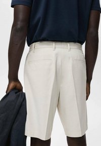 Beiga shorts med skräddarsydd passform, med en slät front, två bakfickor och en midjeband. Modellen håller en marinblå jacka.