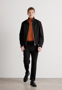 Čierna zipsová bunda cez oranžový rolák, v kombinácii s čiernymi nohavicami a čiernymi topánkami. Hladká textília, minimalistický dizajn, póza pri chôdzi.