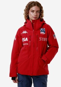 Giacca impermeabile rossa con cappuccio, dotata di due tasche con zip, dettagli con logo e polsini regolabili. Dettagli riflettenti sulla parte anteriore.