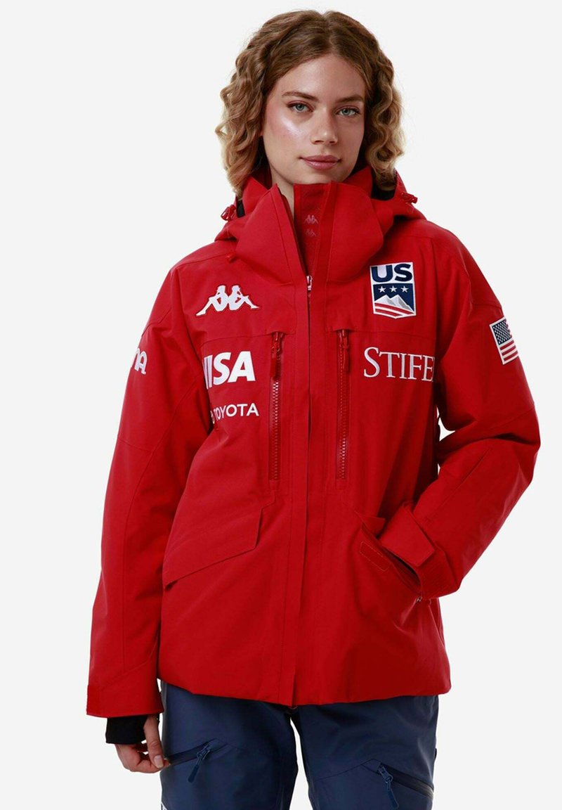 Giacca impermeabile rossa con cappuccio, dotata di due tasche con zip, dettagli con logo e polsini regolabili. Dettagli riflettenti sulla parte anteriore.