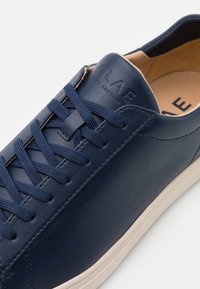 Zapatilla de cuero navy con superficie texturizada, cordones planos y diseño minimalista. Suela de goma color crema y detalle de logo en relieve.