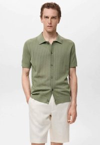 Camisa de botones de manga corta de punto en verde claro con textura de canalé, que presenta un collar y un dobladillo recto, combinada con pantalones cortos beige.
