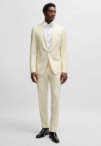 Cream tuxedo jacket with shawl lapels, white button-up shirt, and bow tie. Beige trousers and shiny black dress shoes complete the outfit.