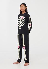 Czarny komplet piżamy z motywem szkieletonu w kremowym kolorze z różowymi akcentami w kształcie serc. Długi rękaw i pasujące spodnie w luźnym fasonie.