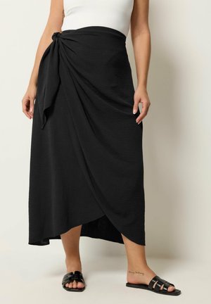 Frau trägt einen schwarzen Wickelrock, der in der Taille gebunden ist, ein weißes Oberteil und schwarze offene flache Sandalen, steht vor einem schlichten hellen Hintergrund.