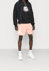 Giacca bomber nera sopra una felpa nera con cappuccio, pantaloni corti pesco con logo Jumpman bianco e sneakers bianche con dettagli blu.