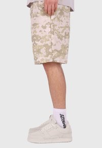 Tarnhosen in Beige-Tönen, mit lockerer Passform, Seitentaschen und besticktem Logo-Detail am Saum; kombiniert mit cremefarbenen Sneakers.