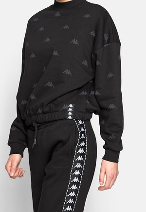 Osoba ubrana w czarną bluzę i spodnie z powtarzającymi się białymi wzorami logo na rękawach, klatce piersiowej i bokach spodni, stojąca na jednolitym tle.