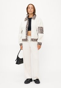 Witte nepbontjas met luipaardprintaccenten, zwarte crop top, crèmekleurige broek en zwarte sneakers. Model houdt een zwarte handtas vast.